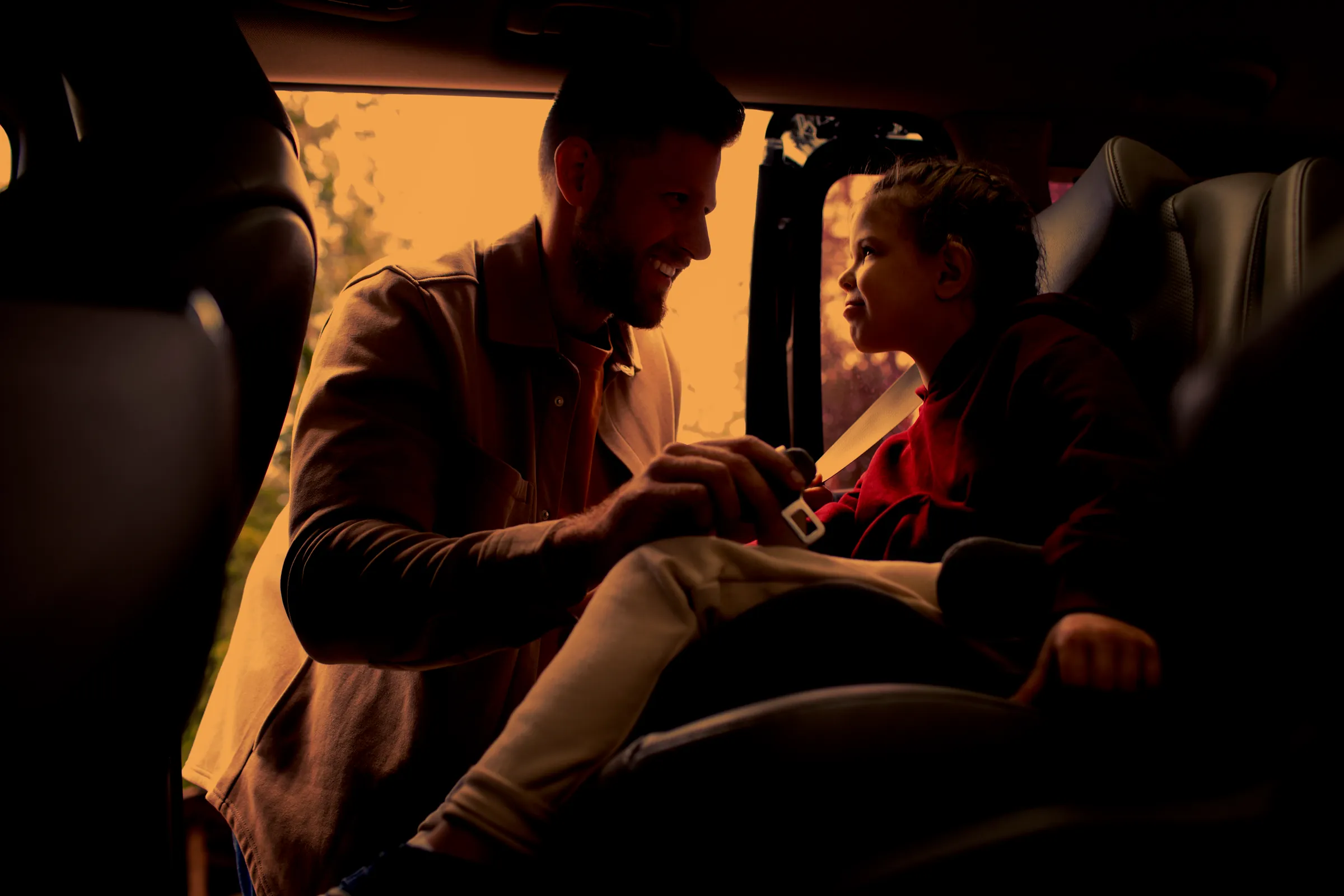A father smiling at his young daughter as he buckles her into a car seat