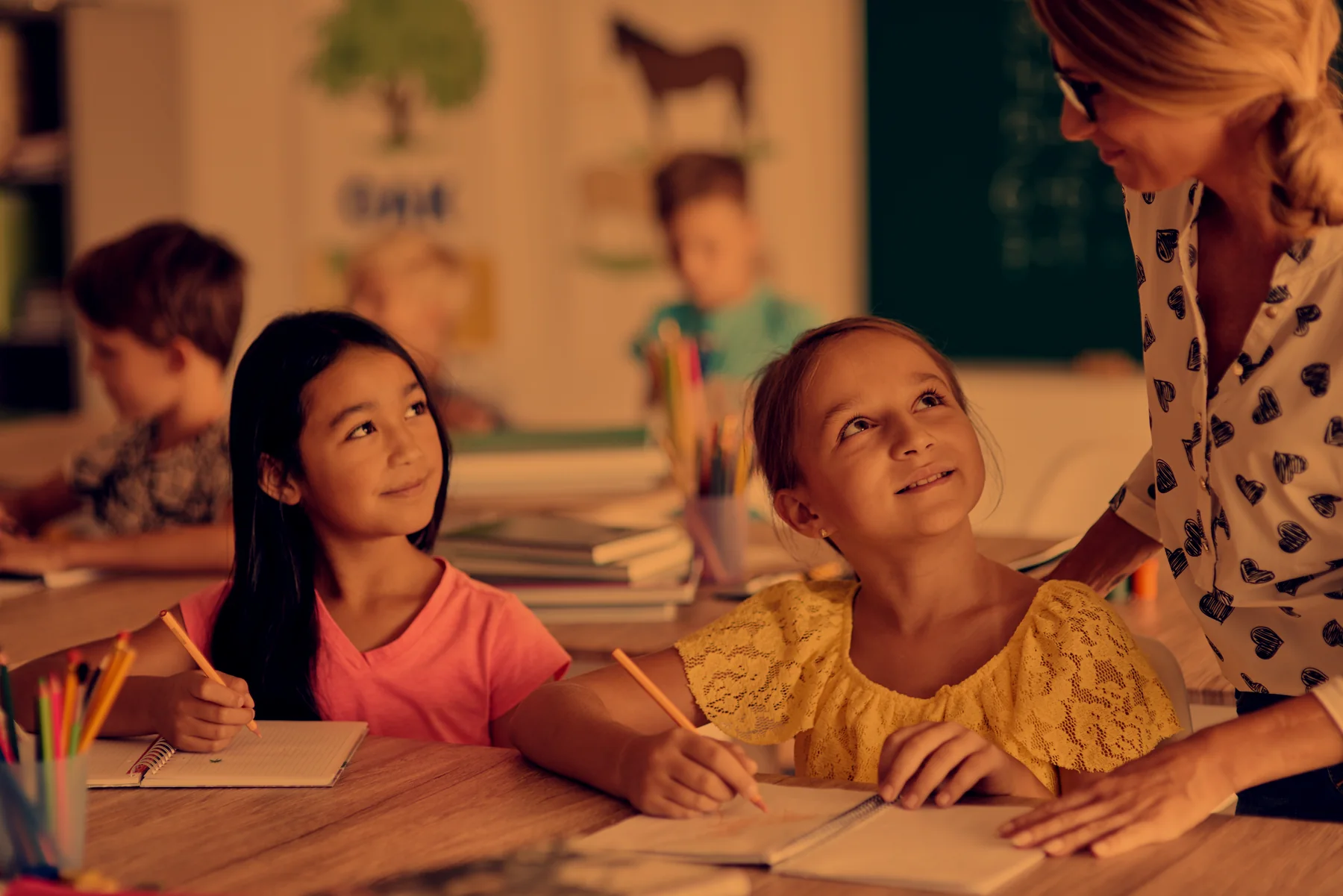 A teacher leaning over two smiling students at their desks in a classroom
